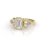 Angled top view showing the emerald cut center and pear side diamonds in a yellow gold bezel setting.
