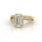 Three-quarter angle view of 3 carat emerald cut bezel set lab diamond ring on yellow gold split shank double band.