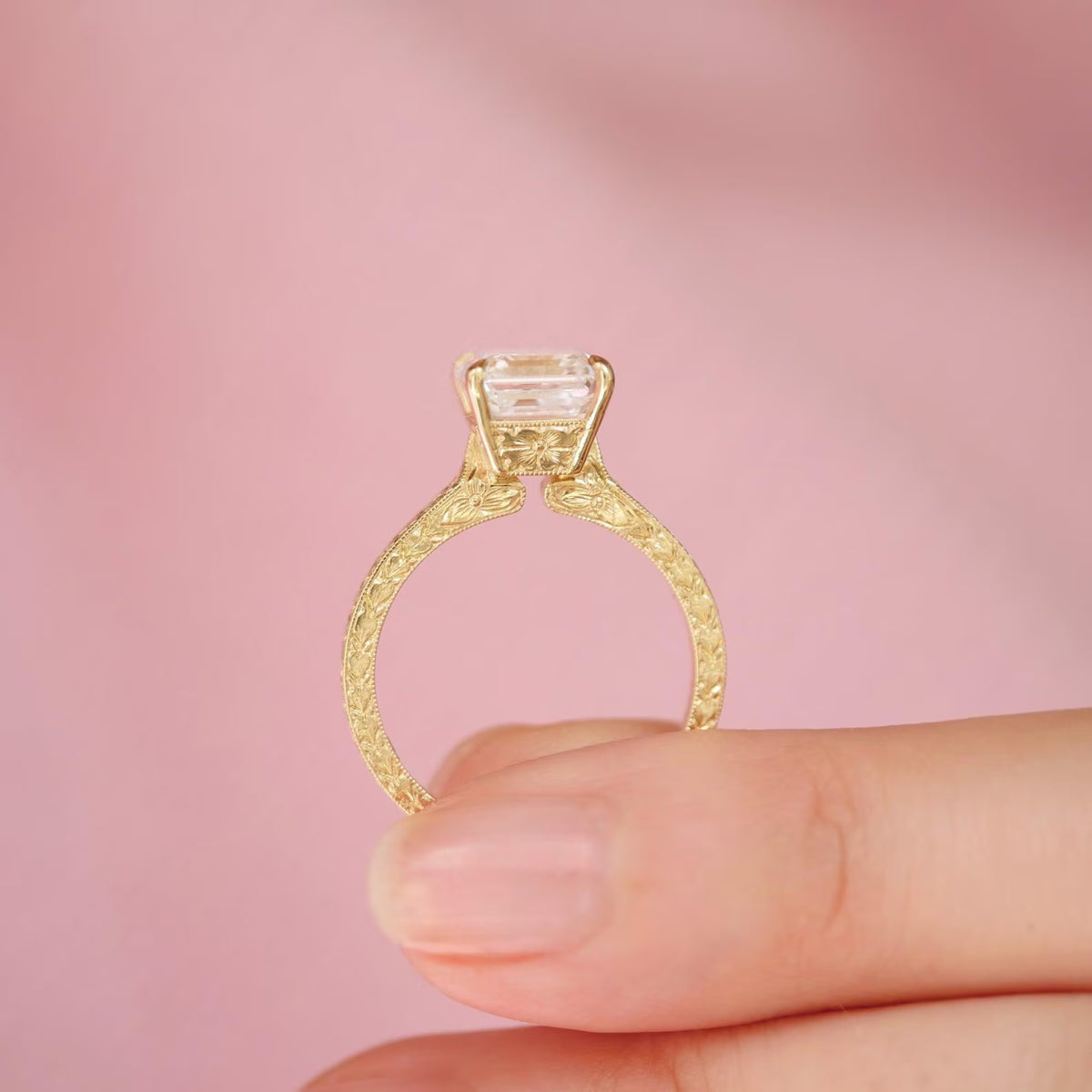Side profile of 2 CT Asscher cut lab grown diamond ring in yellow gold held between fingers showing floral engraved basket gallery