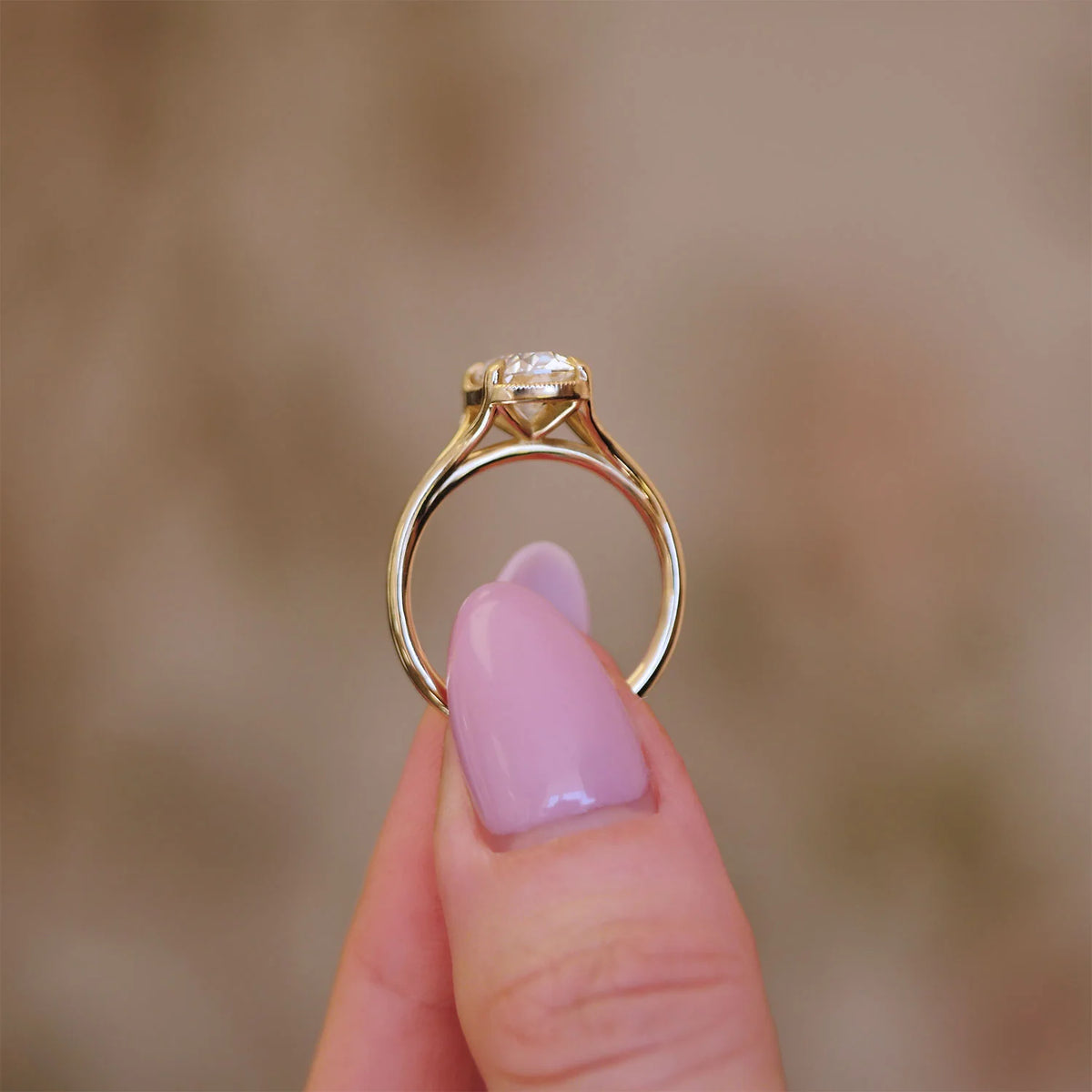 Side profile view of a 3 carat oval lab grown diamond ring in yellow gold held between fingers, featuring a milgrain detailed bezel basket.