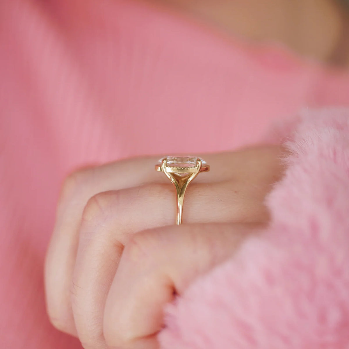 Side perspective view of a 3ct oval lab diamond engagement ring with split shank setting worn on a finger against a pink knit sweater.