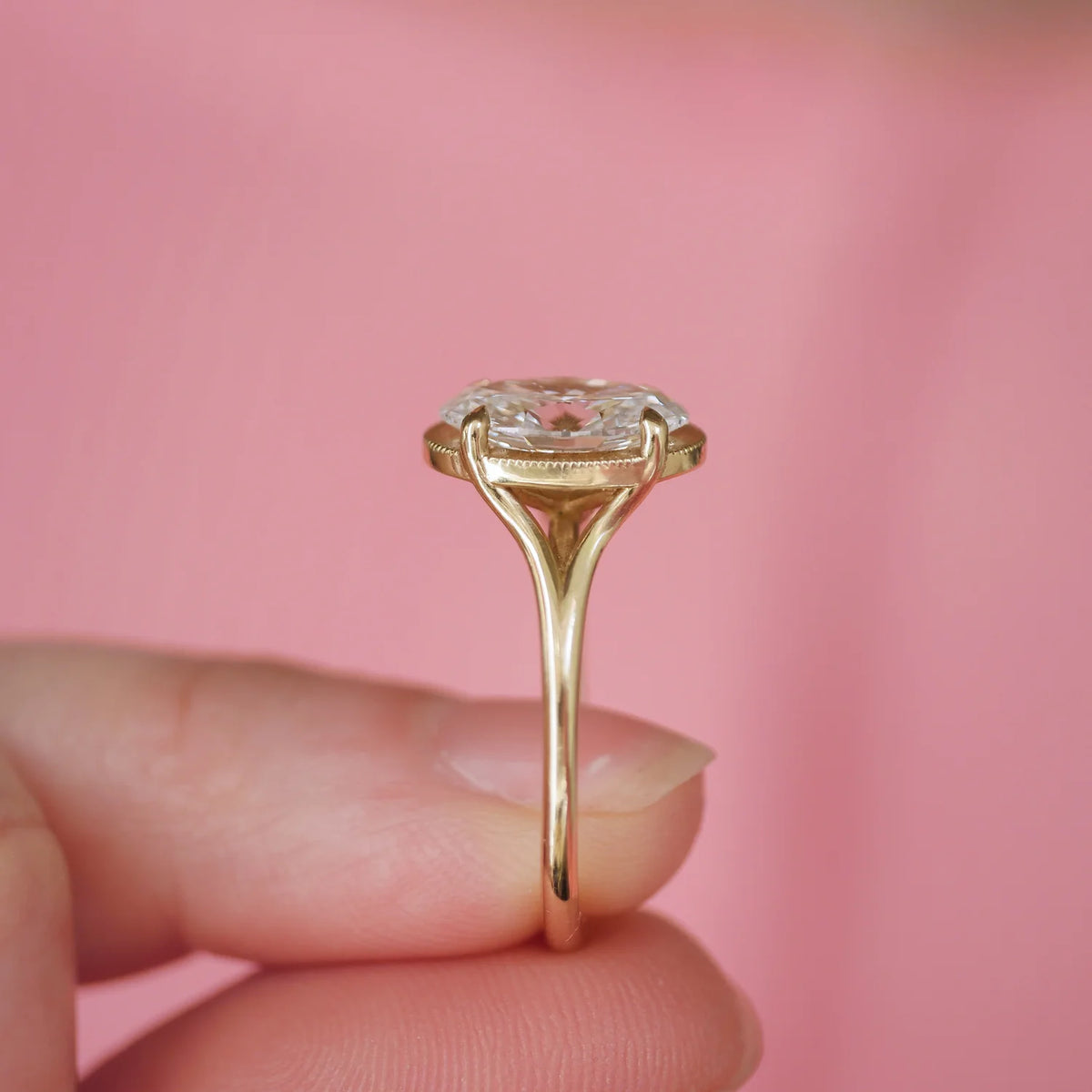 Close-up side profile shot of a 3 carat oval lab diamond ring held against a pink backdrop showing the open undergallery work.