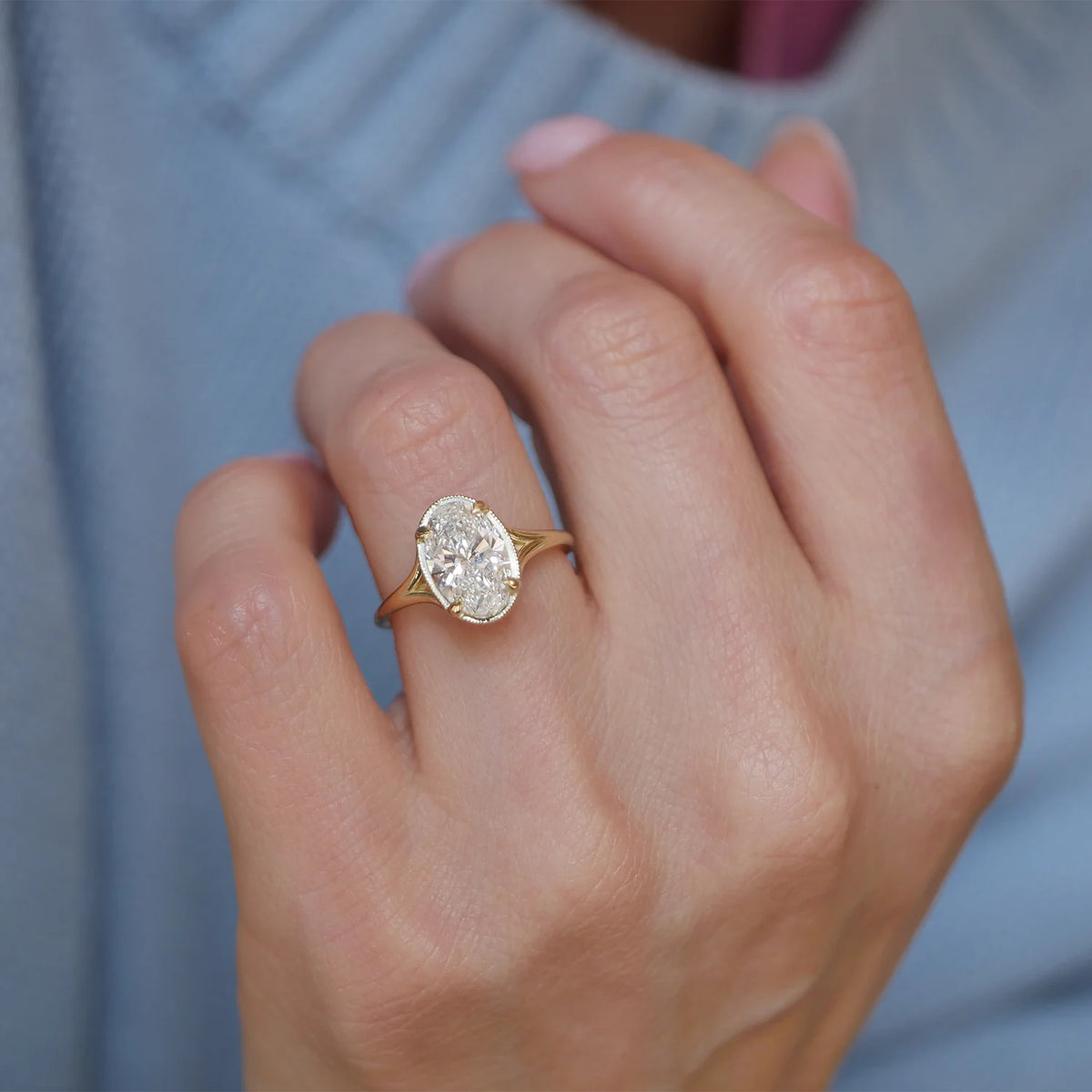 Close-up model hand shot wearing the 3 carat oval lab diamond milgrain bezel ring in yellow gold with blue sweater sleeve.