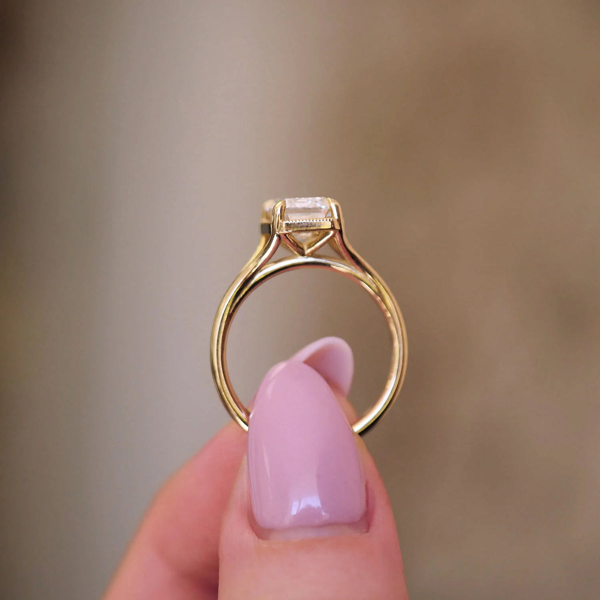 Side profile view of a 3 carat emerald cut lab grown diamond ring held between fingers, showing the milgrain bezel basket and cathedral setting.