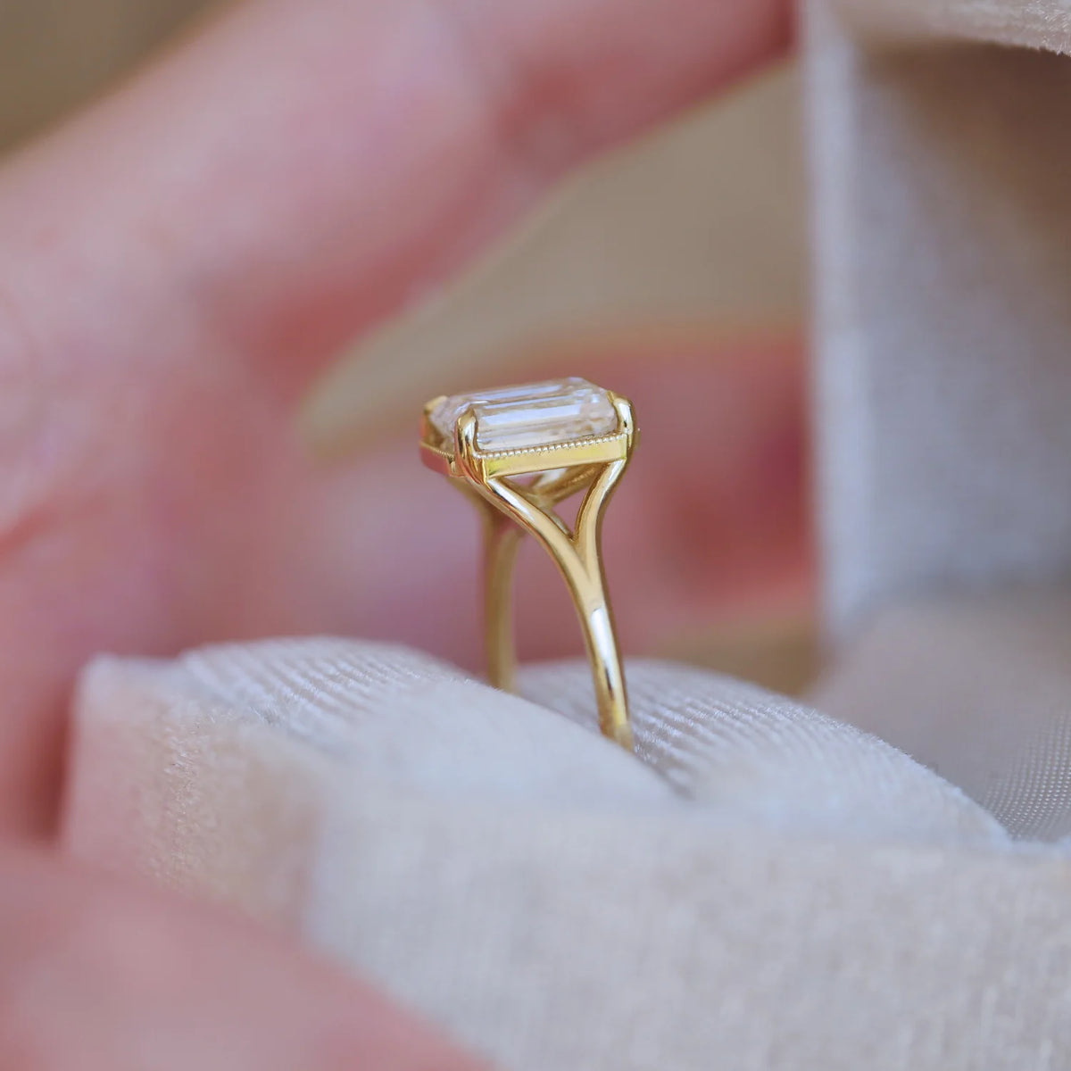 Profile perspective of a 3 carat emerald cut lab diamond split shank engagement ring resting in a white velvet jewelry box.