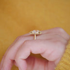 A gold ring with a large cushion-cut clear gemstone in a halo setting, worn on a person's finger against a warm yellow background.
