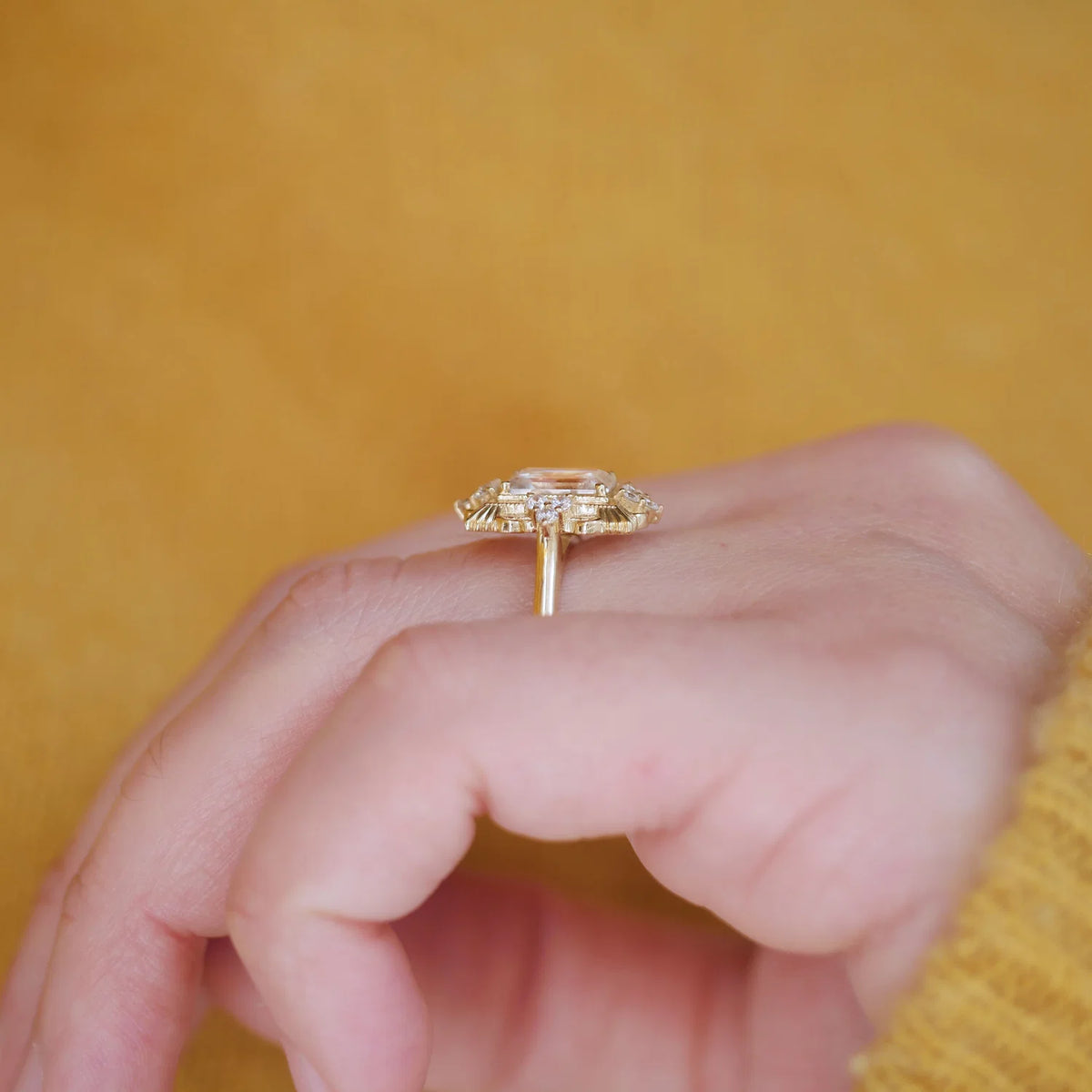 A gold ring with a large cushion-cut clear gemstone in a halo setting, worn on a person's finger against a warm yellow background.
