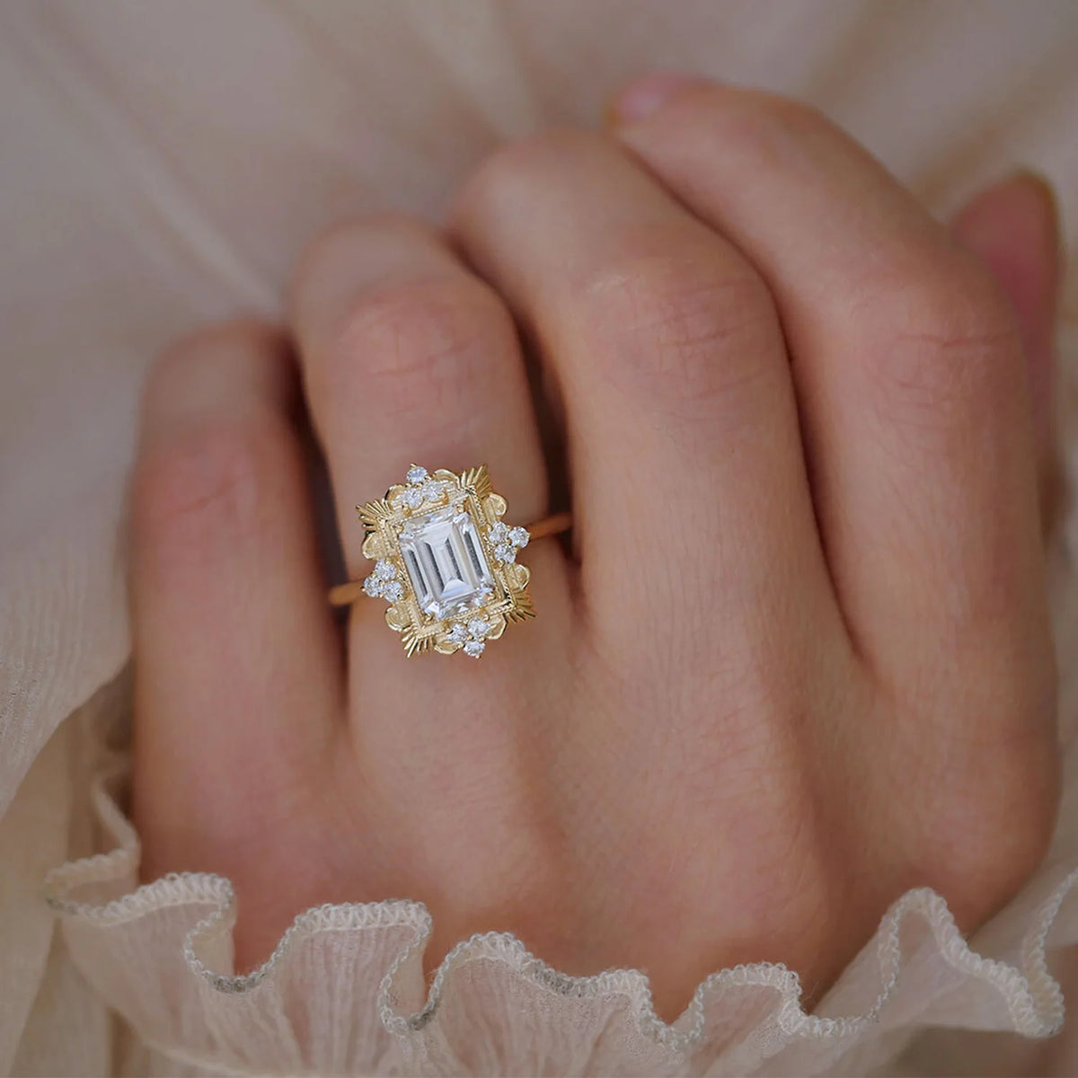 Lifestyle shot of a 3 carat emerald cut lab diamond ornate halo ring worn on a finger paired with a sheer ruffled sleeve.