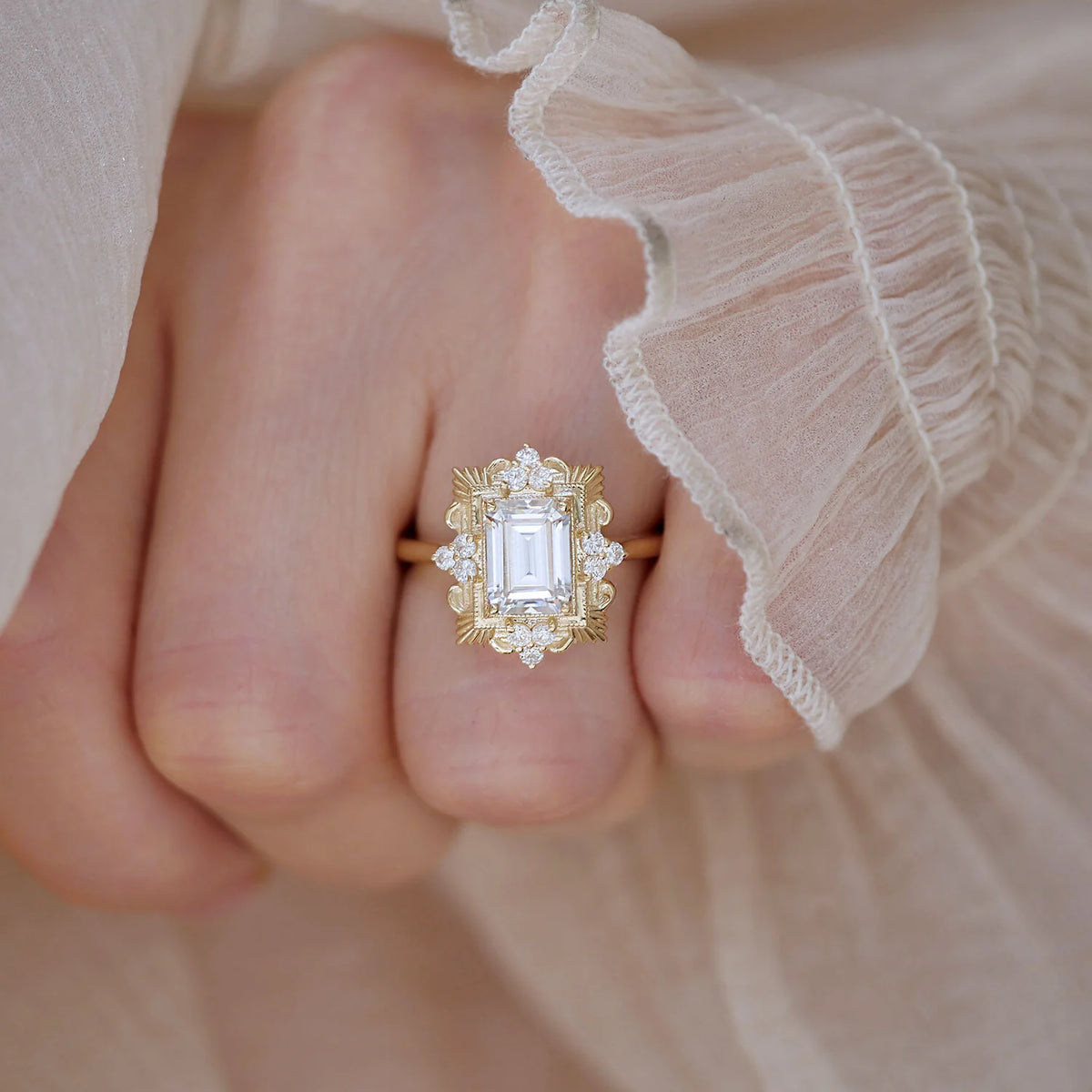 Top-down hand shot of a woman wearing a 3 carat emerald cut lab diamond ornate halo ring against romantic ruffled silk cuff.