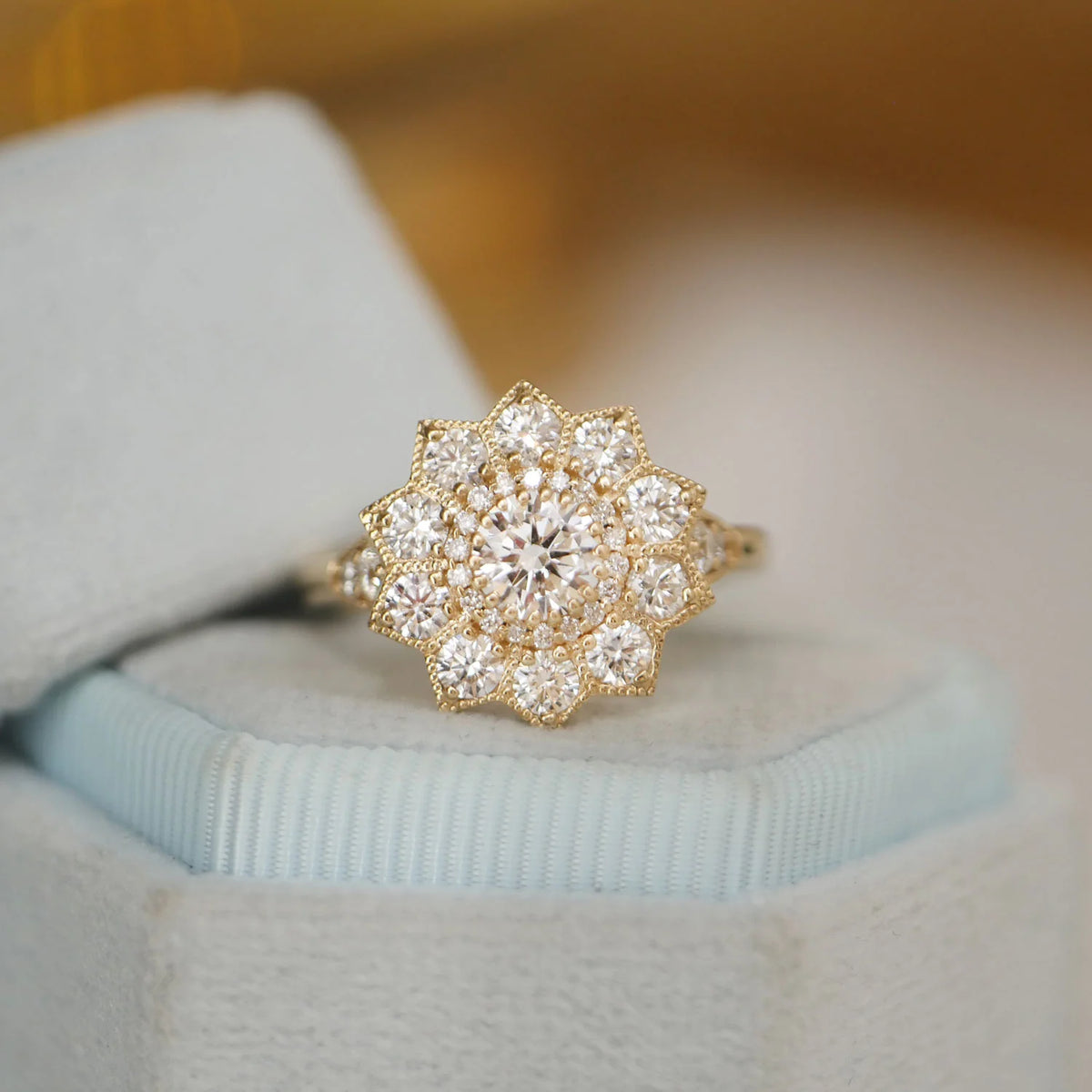 Angled view of a 1.5 CT round lab grown diamond starburst floral halo engagement ring in yellow gold placed inside a light blue velvet ring box.