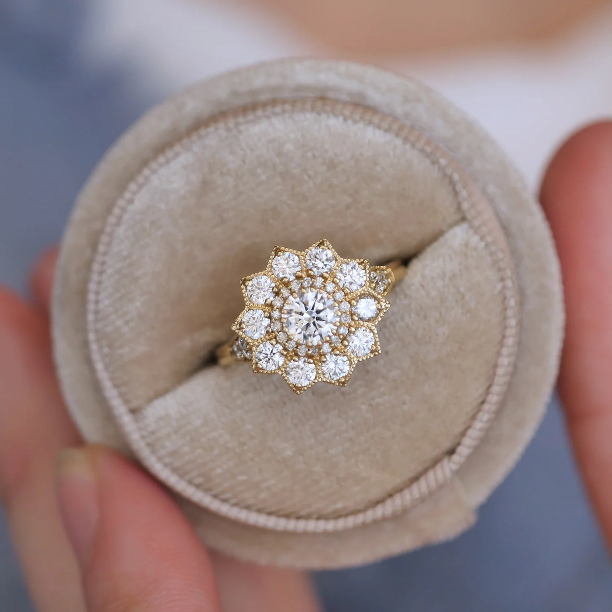 Overhead view of a 1.5 carat round lab created diamond floral halo ring seated in a round beige velvet jewelry box.