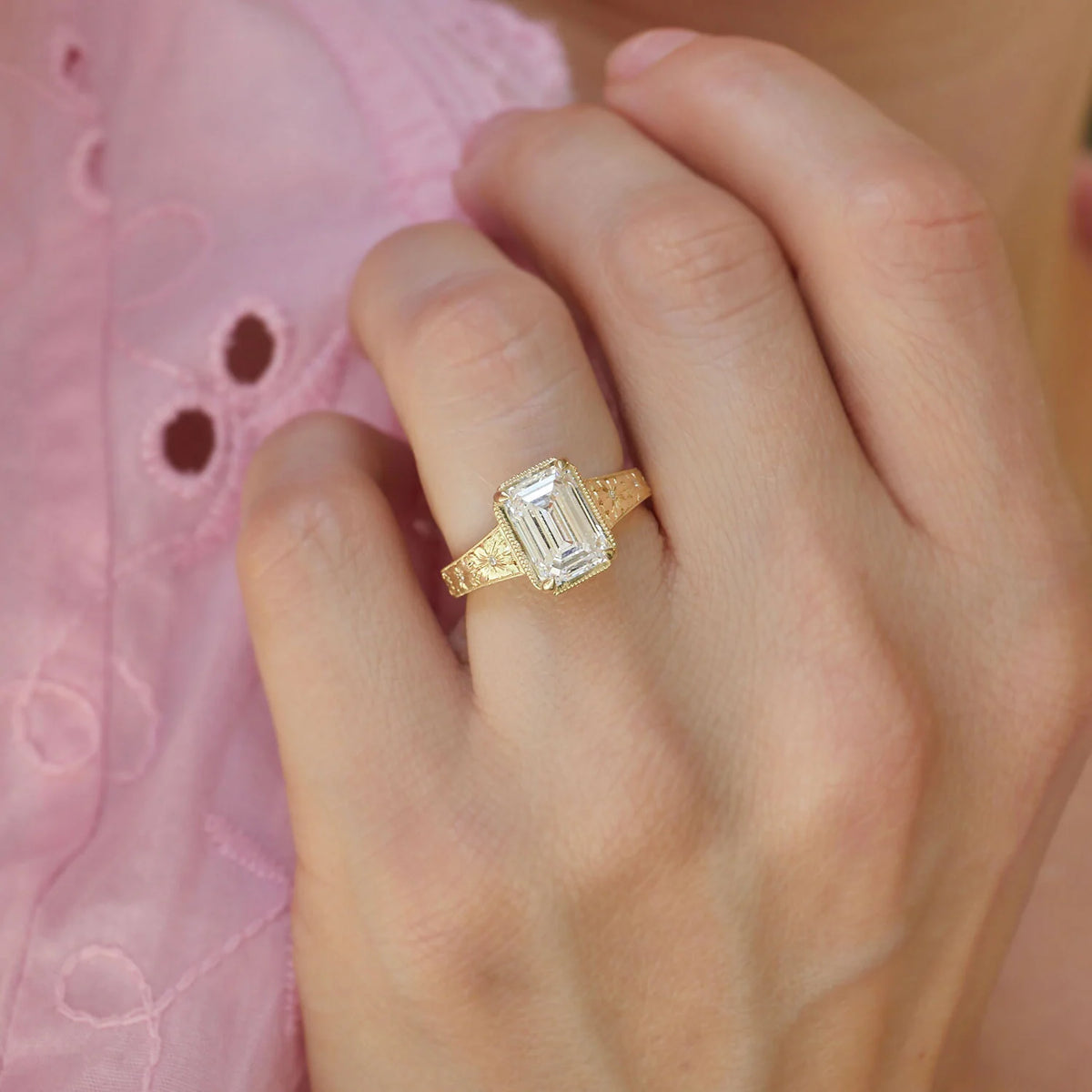 On-finger view of a 3 CT emerald cut lab grown diamond engagement ring in yellow gold featuring a floral engraved bezel band.