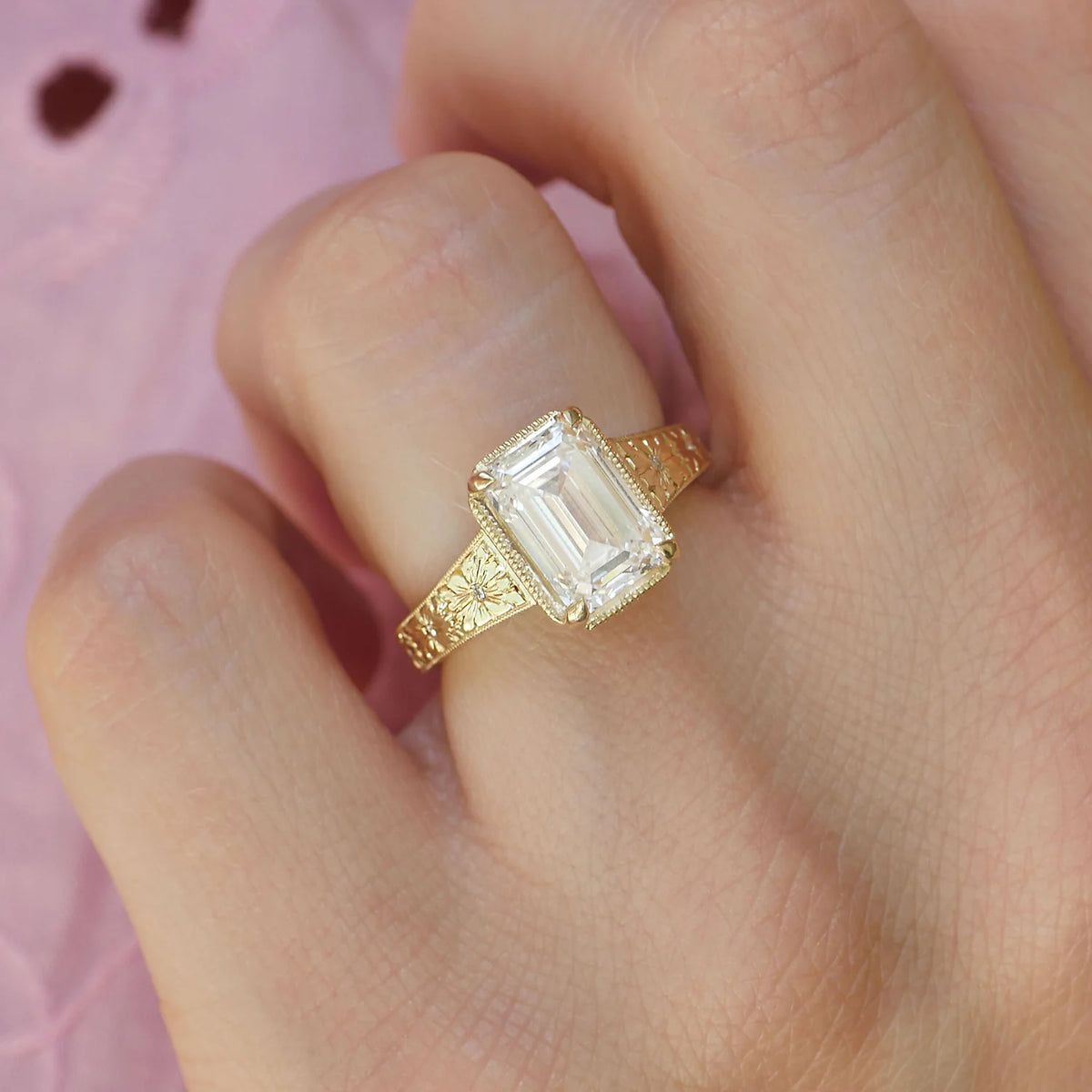 Angled top-down hand view showing a 3 carat emerald cut lab created diamond ring with vintage starburst floral engraving.
