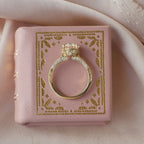 Side profile view of a 2 carat round lab grown diamond filigree engagement ring sitting on a pink leather vintage book.