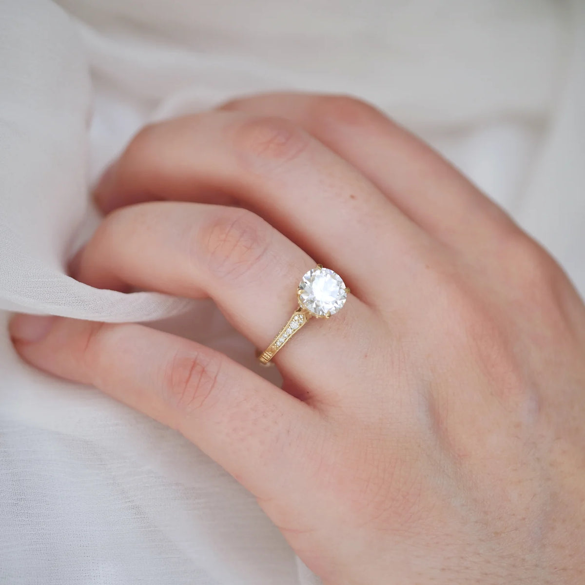 A close-up of a hand wearing a yellow gold engagement ring with a round brilliant-cut clear center stone and a pavé band, resting against soft white fabric.