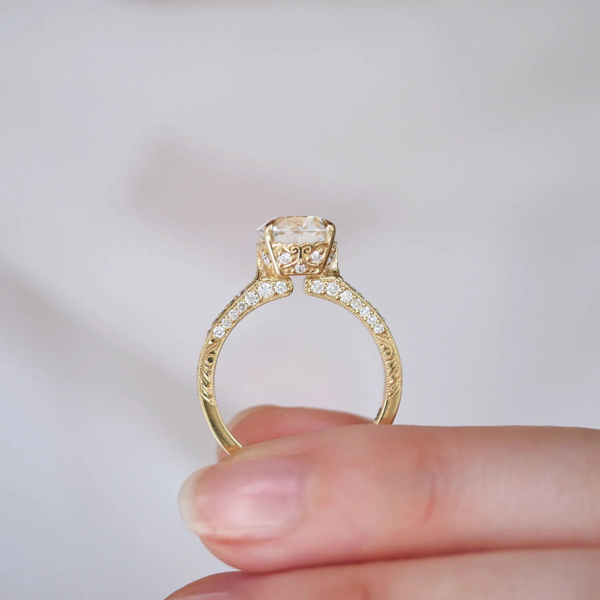 Hand holding a yellow gold 2 CT round lab diamond ring showing the side profile scrollwork and hidden accent diamonds.