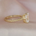 Side view of a yellow gold 3 carat emerald lab diamond ring resting on pink silk, displaying the ornate etched gallery and band details.