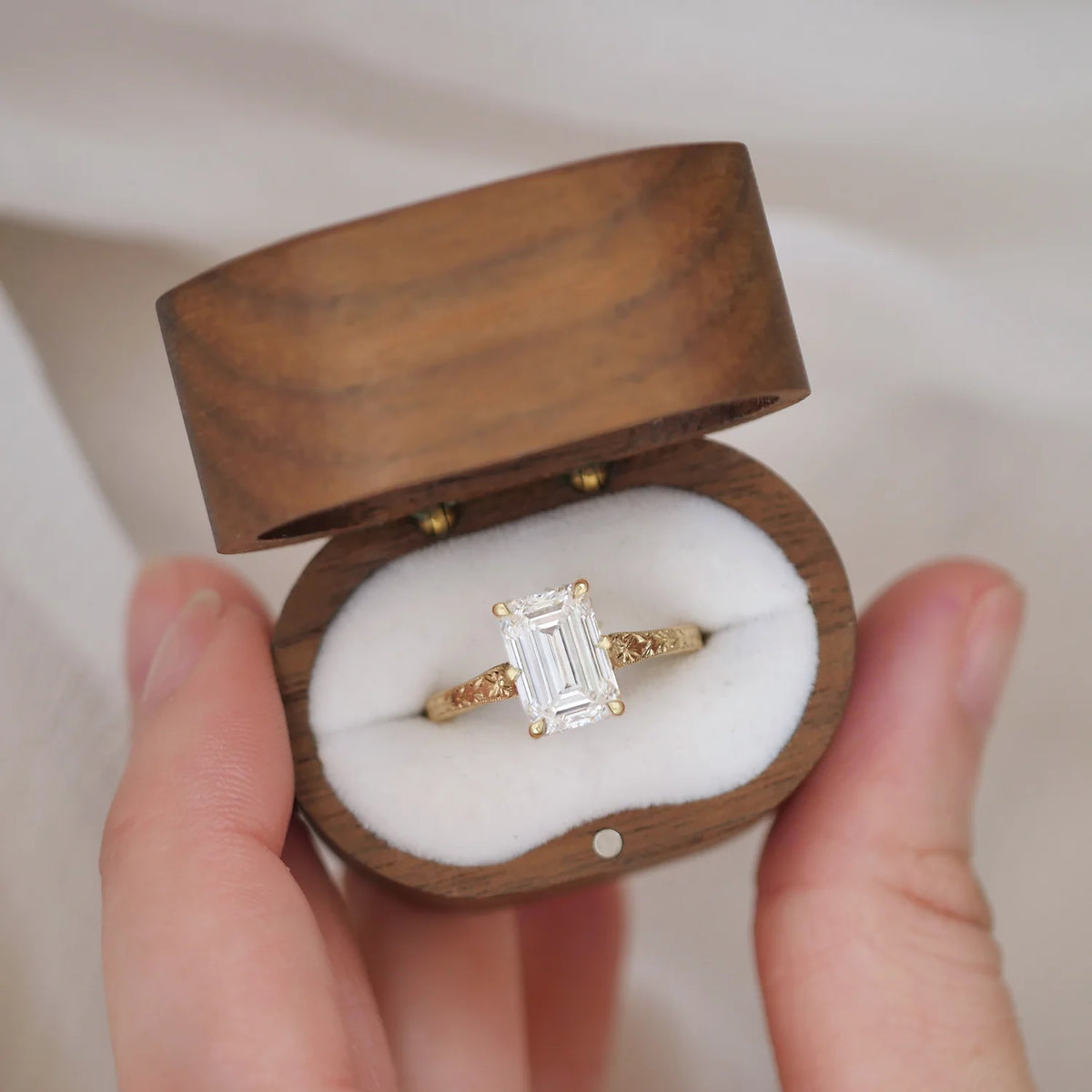 Hand holding an open oval wooden ring box displaying a 3 CT emerald cut lab grown diamond engagement ring with a yellow gold carved band.