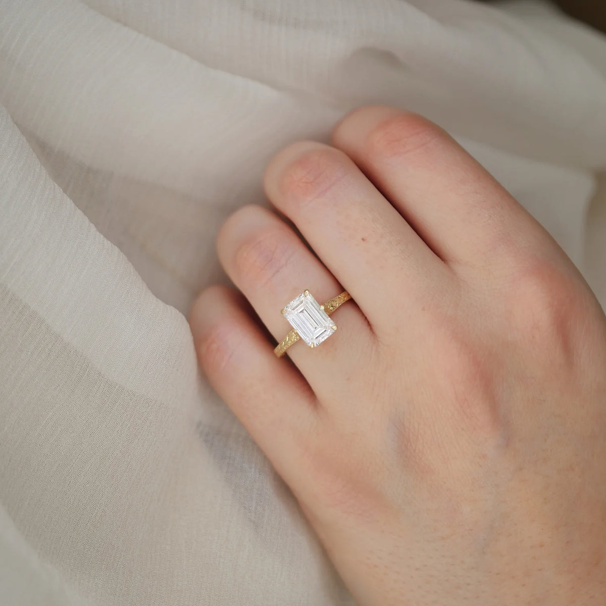 On-hand photo showing a 3 CT emerald cut lab grown diamond vintage engraved solitaire ring worn on finger over fabric.