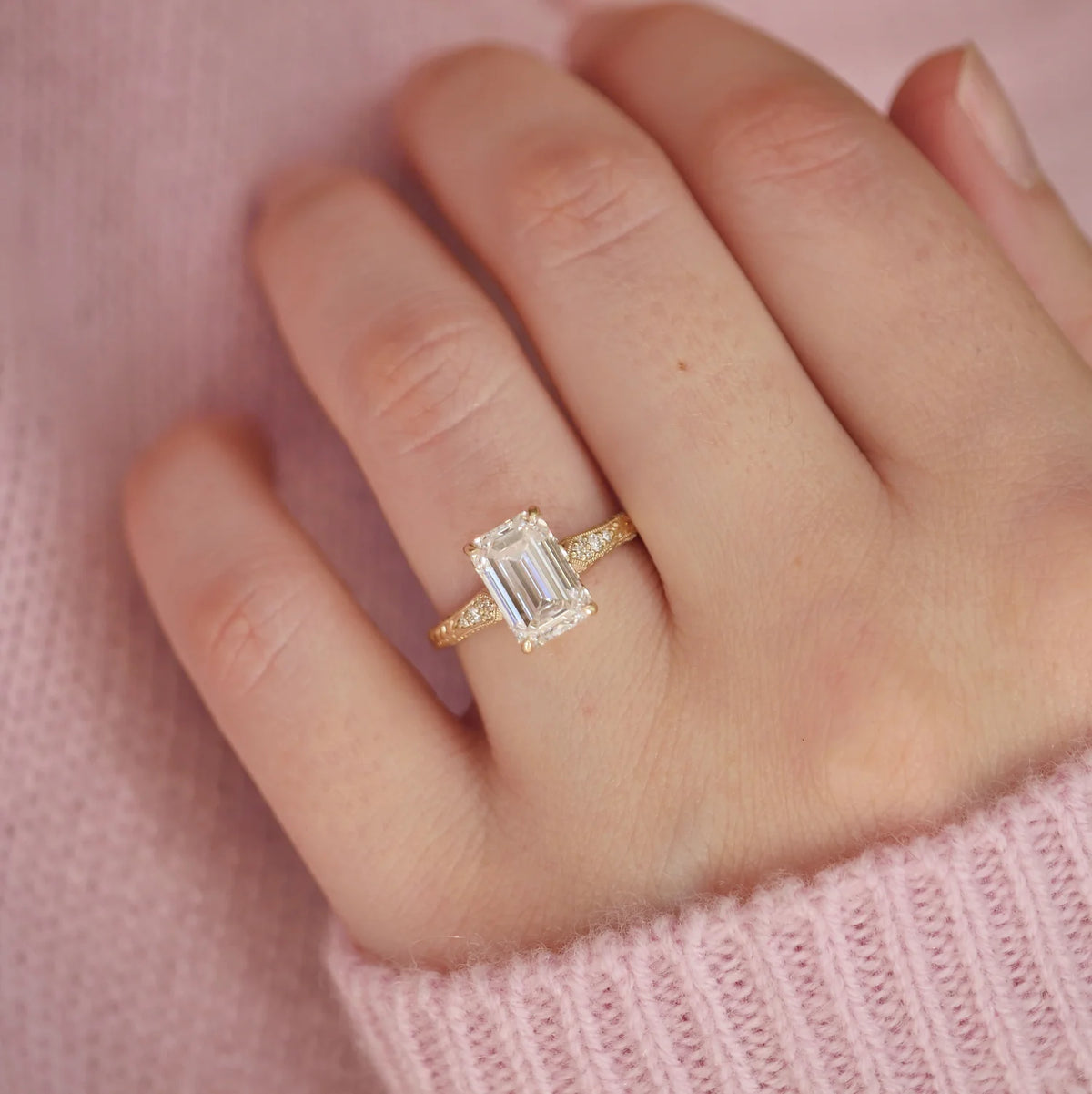 On-hand view of a 3 CT emerald cut lab diamond ring featuring engraved botanical patterns and sparkling side accent diamonds against a pink sweater background.