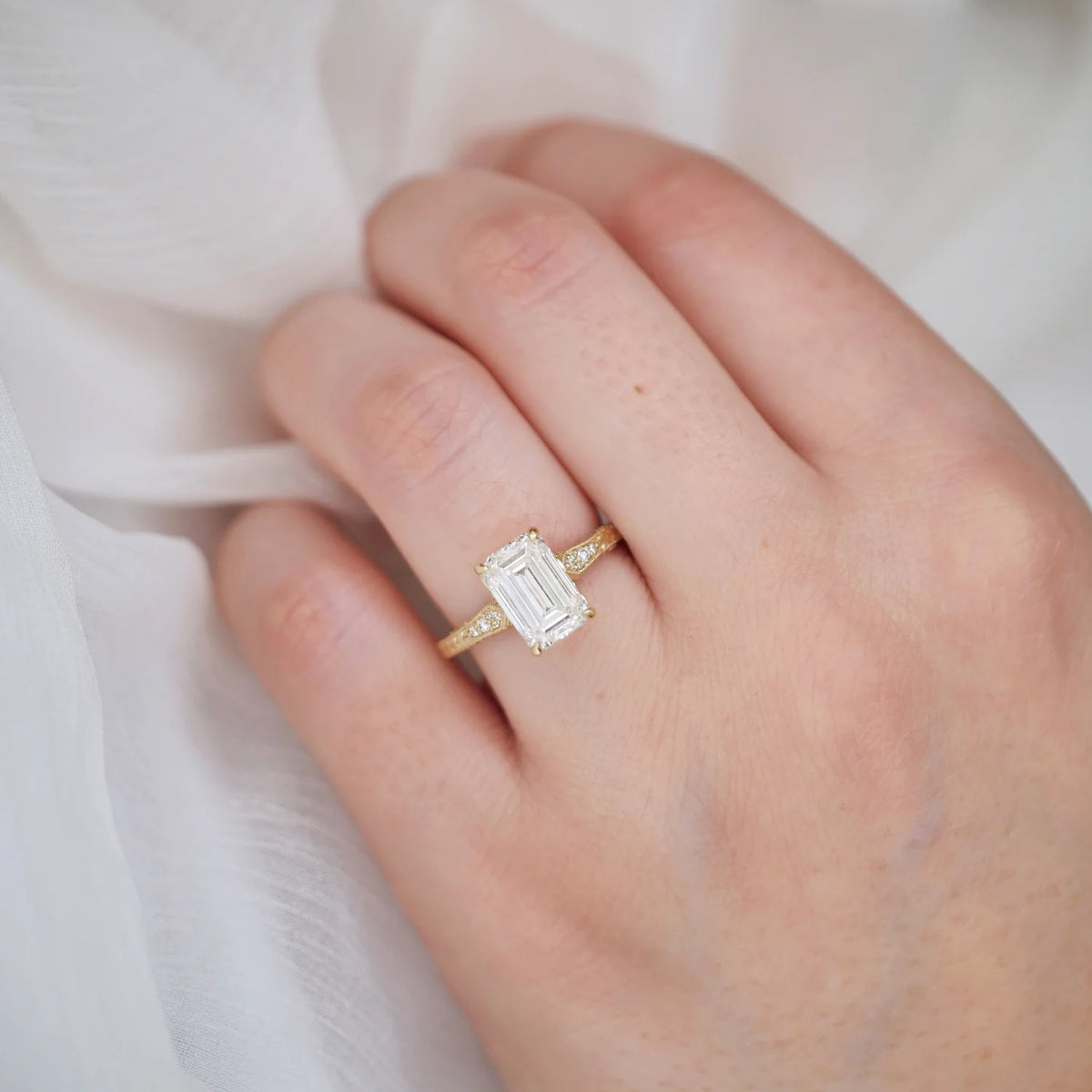 3 CT emerald cut lab grown diamond solitaire with side diamond accents and hand-engraved detailing worn on a hand over soft white fabric.
