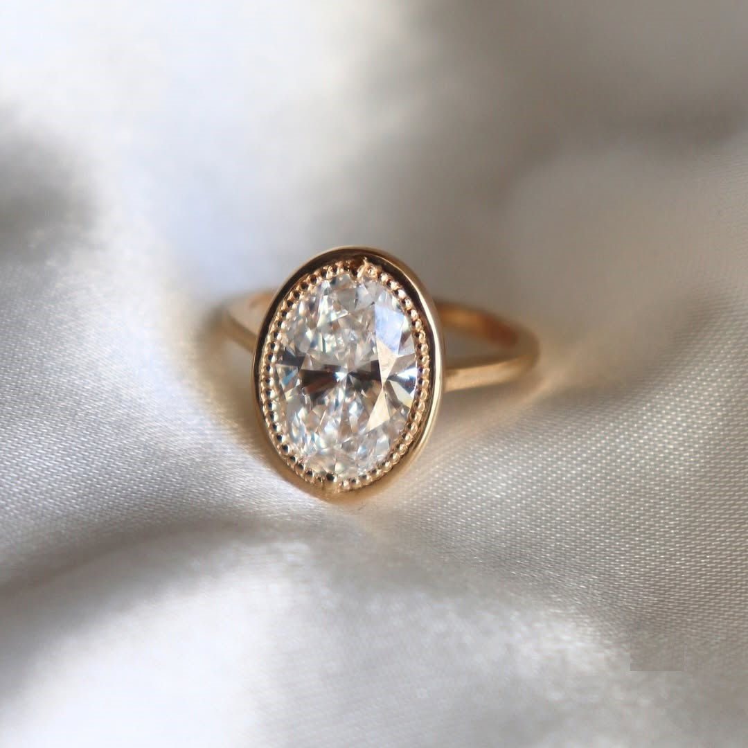 Front close-up view of a 3 carat oval cut lab grown diamond with a vintage milgrain bezel rim on a polished yellow gold band.