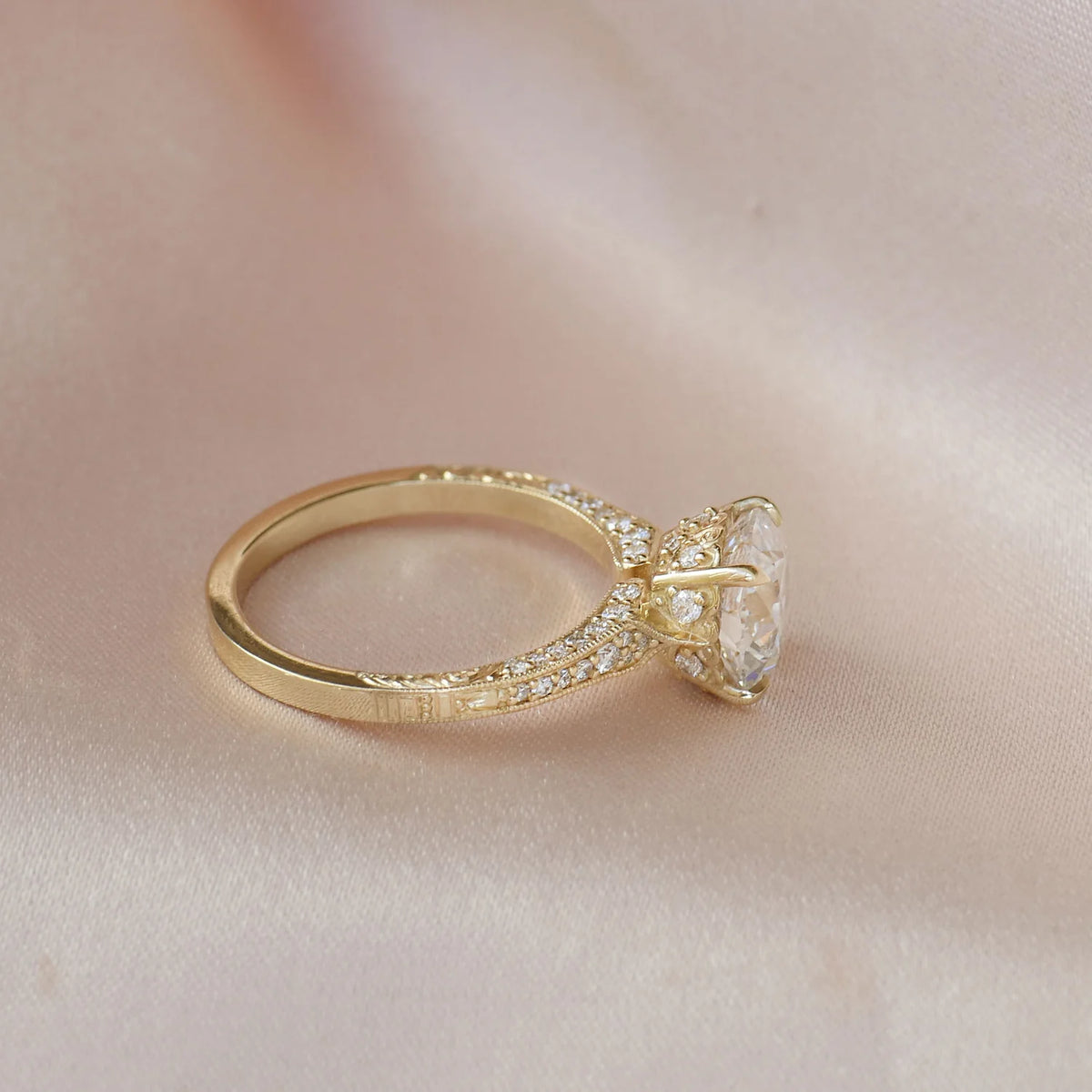 Side profile shot of a yellow gold 2 CT oval diamond ring with ornate under-gallery resting on silk.