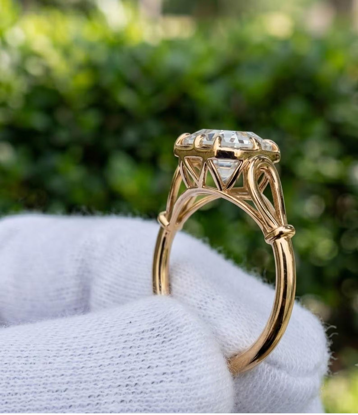 A yellow gold ring with a clear asscher-cut gemstone held on a white textured glove, set against a blurred green outdoor background.