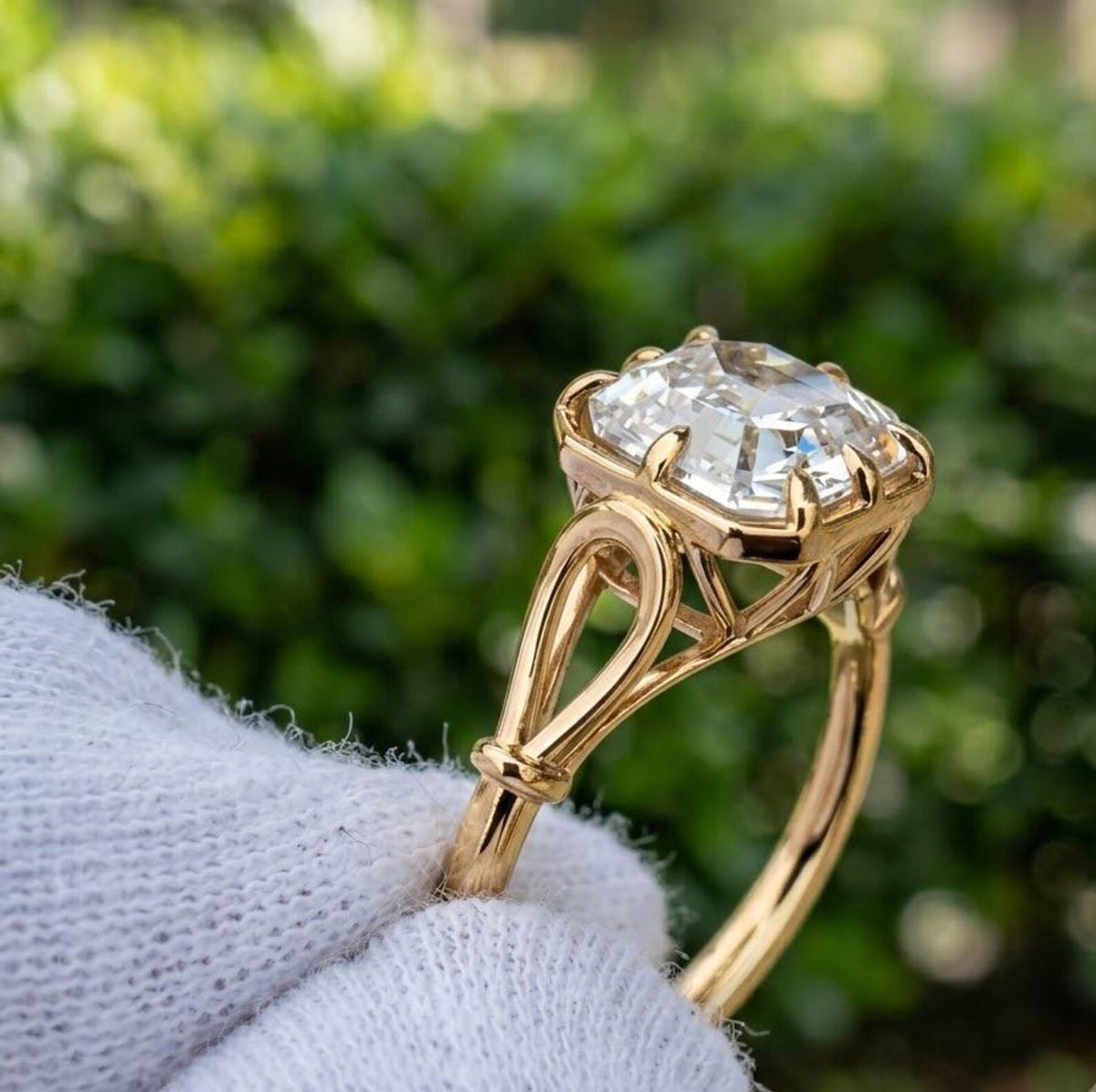 A yellow gold ring with a asscher-cut clear gemstone held by a white gloved hand, set against a blurred green garden background.