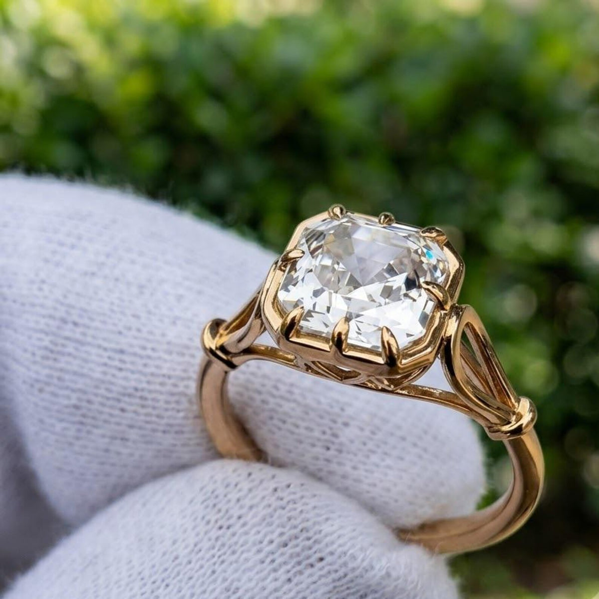 A gold ring with a large clear asscher-cut gemstone, held on a white gloved finger against a blurred green outdoor background.