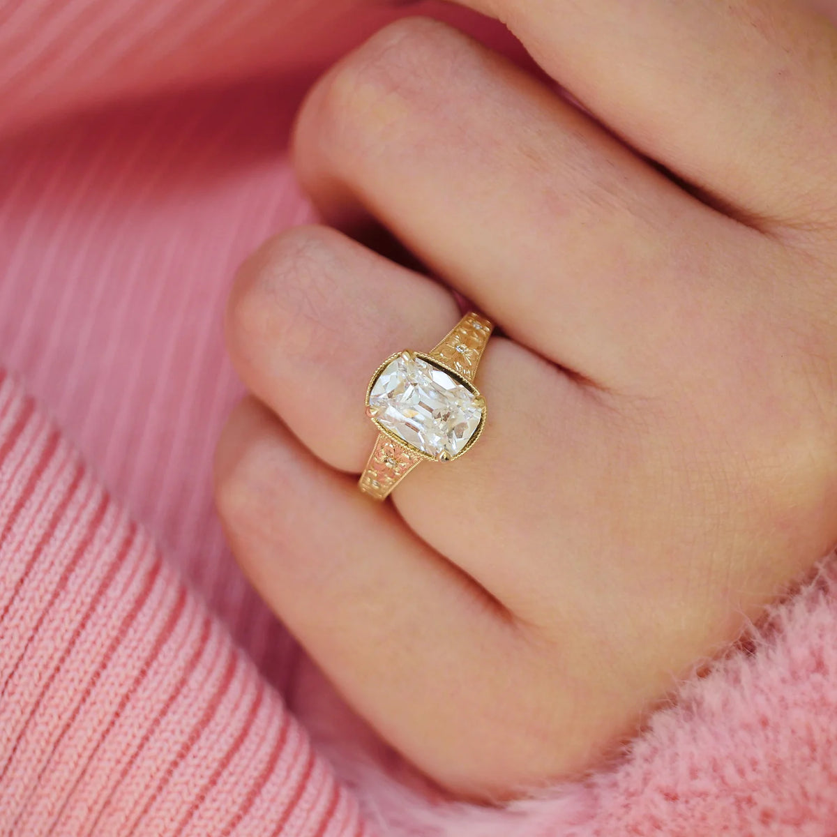 Close-up of a 3 carat cushion cut lab diamond ring with milgrain bezel and floral engraved shank worn on a finger.