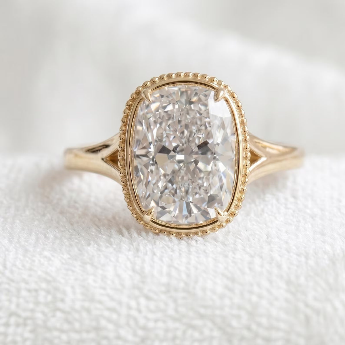 Top-down view of a 3 CT elongated cushion cut lab grown diamond engagement ring with a beaded milgrain bezel on white fabric.