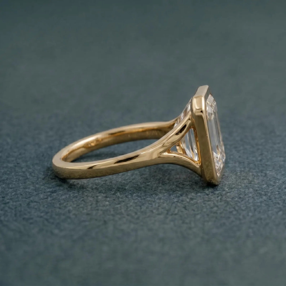 Side profile view of a 3 CT emerald cut lab grown diamond ring featuring a smooth yellow gold bezel setting and split-shoulder band.