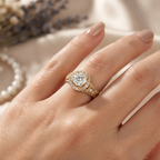 A close-up of a hand wearing a gold ring with a large round clear center stone and small surrounding stones, photographed against a soft cream fabric background with blurred dried flowers and a pearl strand.