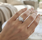 A heart-shaped diamond ring with a halo setting on a hand with manicured nails, photographed in a softly blurred indoor setting.