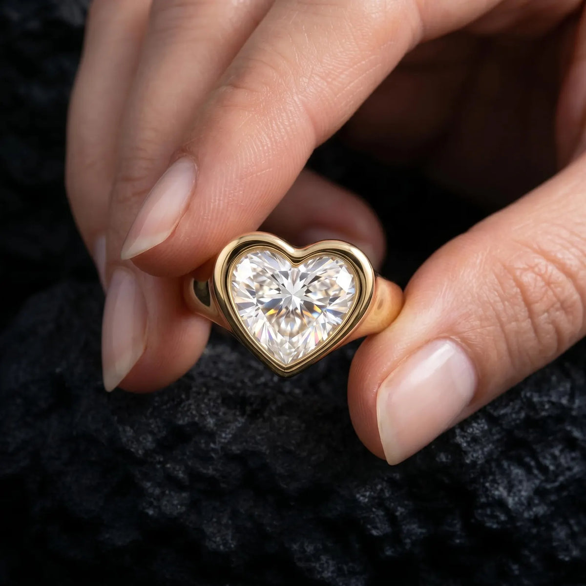Hand holding a 3 CT heart-shaped lab-grown diamond bezel ring against a textured dark background.