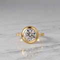 3 CT round lab grown diamond vintage bezel ring in yellow gold resting on a white marble surface displaying center stone reflections.