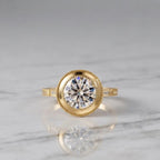 3 CT round lab grown diamond vintage bezel ring in yellow gold resting on a white marble surface displaying center stone reflections.