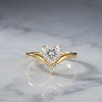 2 carat heart-shaped lab diamond chevron engagement ring in yellow gold set on a reflective white marble surface.