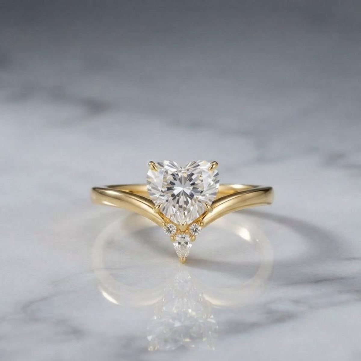 2 carat heart-shaped lab diamond chevron engagement ring in yellow gold set on a reflective white marble surface.