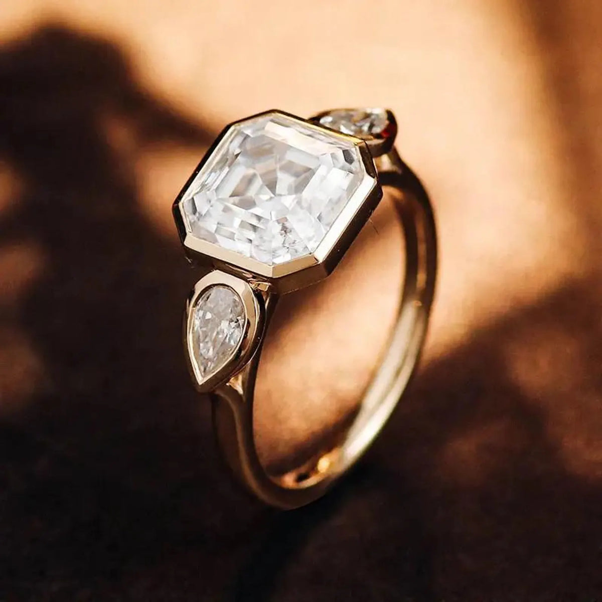 Angled view of a 3 CT Asscher and pear cut lab diamond three-stone bezel setting under warm lighting.