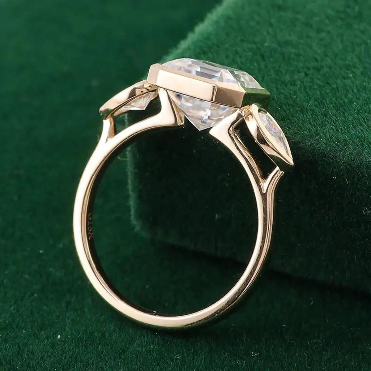 Side view of a 18K yellow gold 3 carat Asscher lab diamond bezel ring resting on dark green velvet background.
