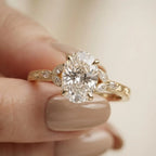 Detailed close-up view of a hand holding a 4 CT oval lab diamond solitaire ring with leaf motif side stones and engraved band.