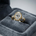 3 CT oval lab grown diamond engagement ring with milgrain bezel and pear side stones in yellow gold presented in a gray velvet box.