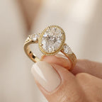 Detailed close-up view of a hand holding a 3 CT oval lab diamond ring displaying pear side accents and botanical hand engraving on yellow gold.