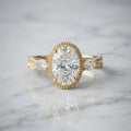 Front view of a 3 CT oval cut lab grown diamond engagement ring on white marble showing milgrain bezel setting and side pear diamonds.