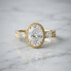 Front view of a 3 CT oval cut lab grown diamond engagement ring on white marble showing milgrain bezel setting and side pear diamonds.