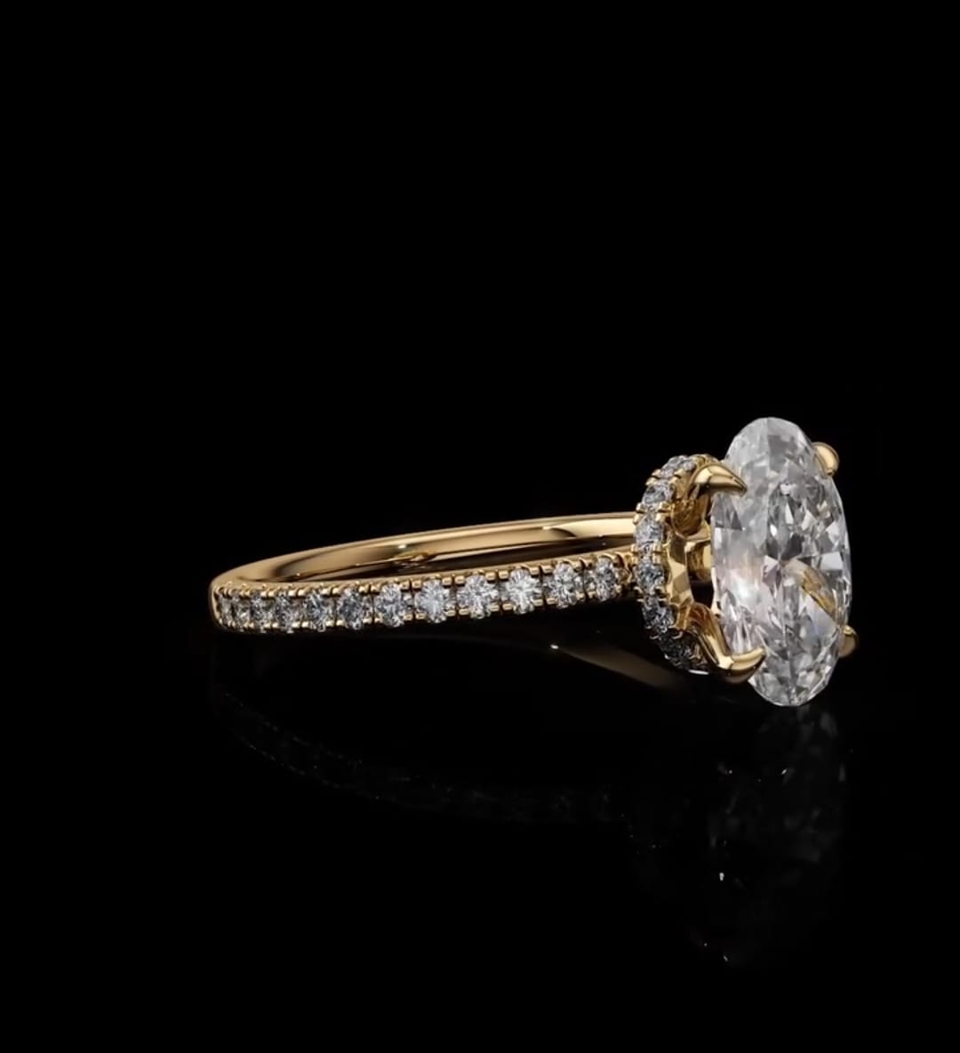 A yellow gold ring featuring a large oval-cut clear center stone in a prong setting, with a halo of small stones and a pavé-set band, shown on a reflective black background.