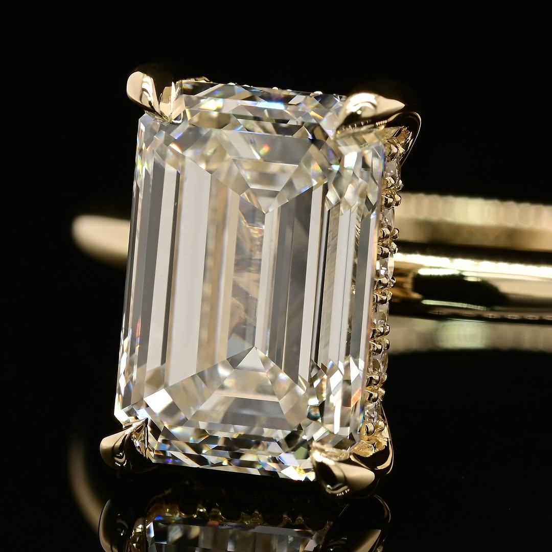 An emerald-cut diamond ring in a yellow gold setting, displayed on a reflective black surface against a dark background.