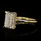 An emerald-cut diamond ring set in yellow gold, featuring a halo of small diamonds along the side of the main stone, shown on a reflective black background.